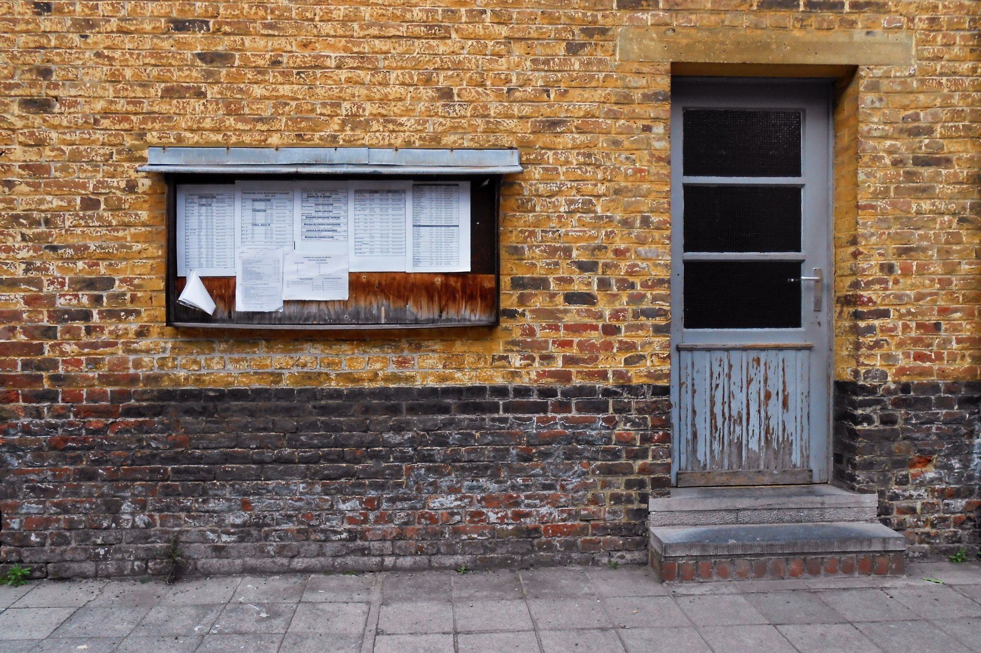 Avisos impresos en una vitrina sobre una pared de ladrillo junto a una puerta gastada, con una hoja despegándose.
